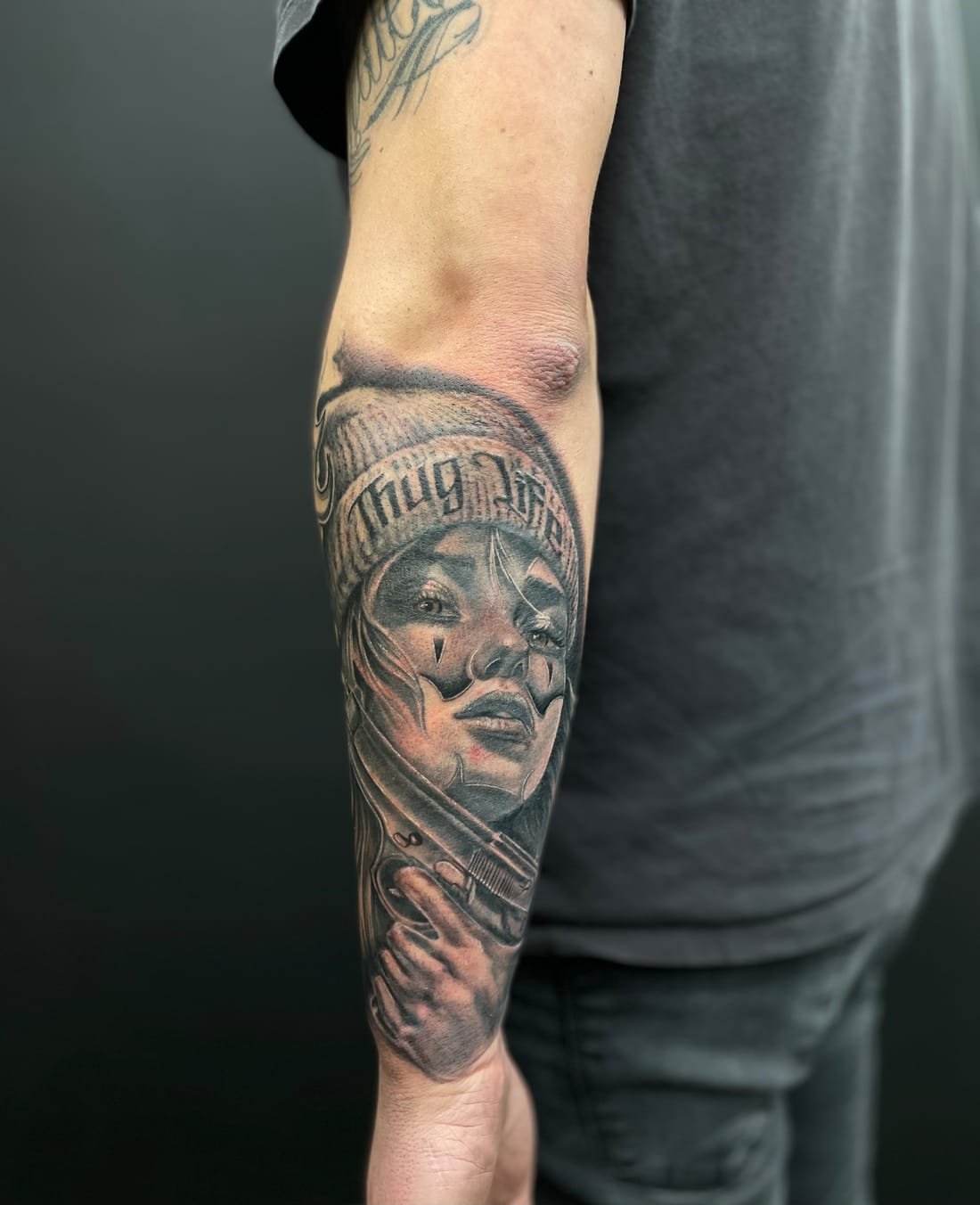 Chicana au bonnet — portrait réaliste noir et gris sur avant-bras, fond sombre