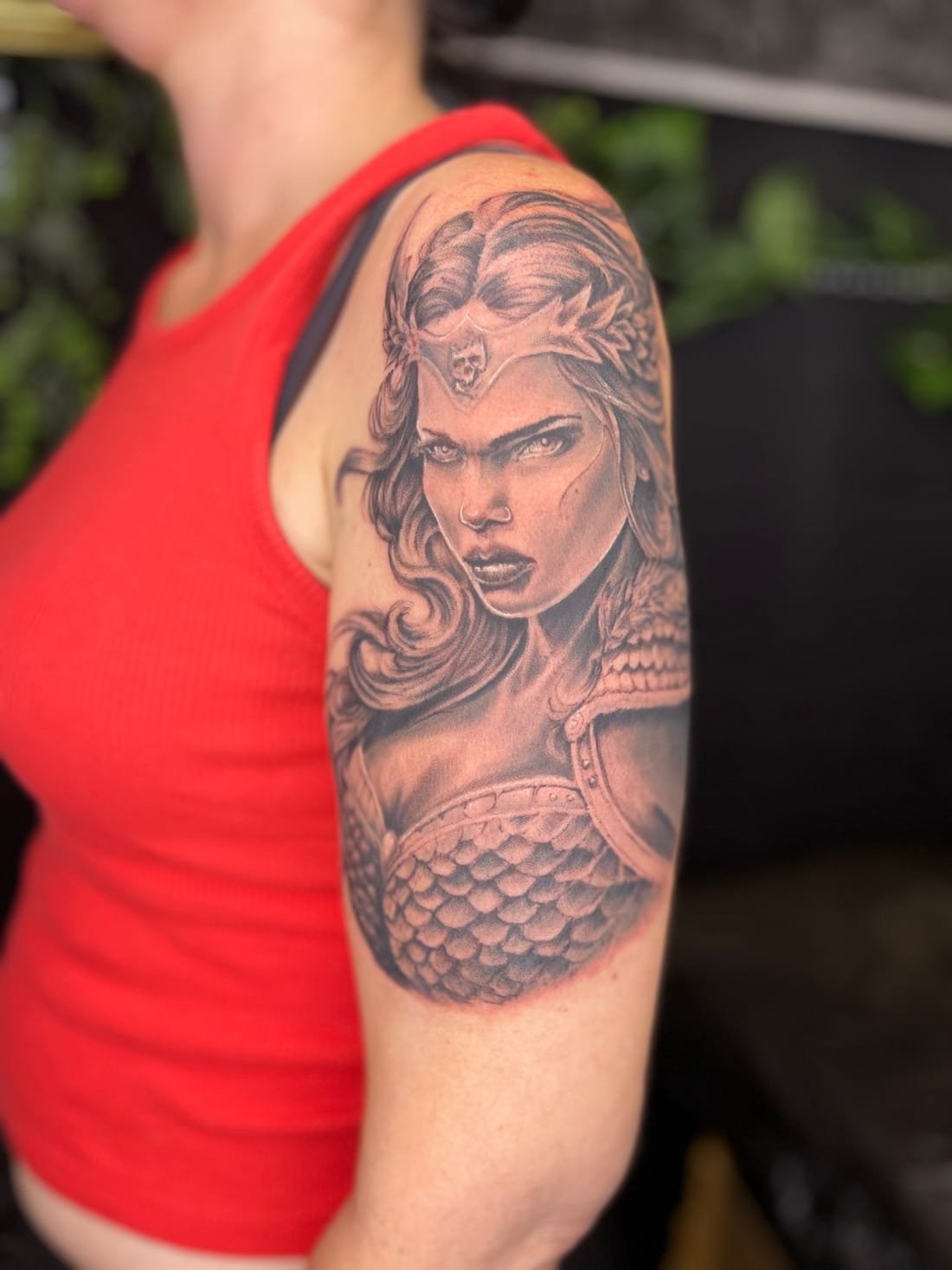 Guerrière amazone — portrait casqué et armure d'écailles, tatouage réaliste noir et gris sur épaule