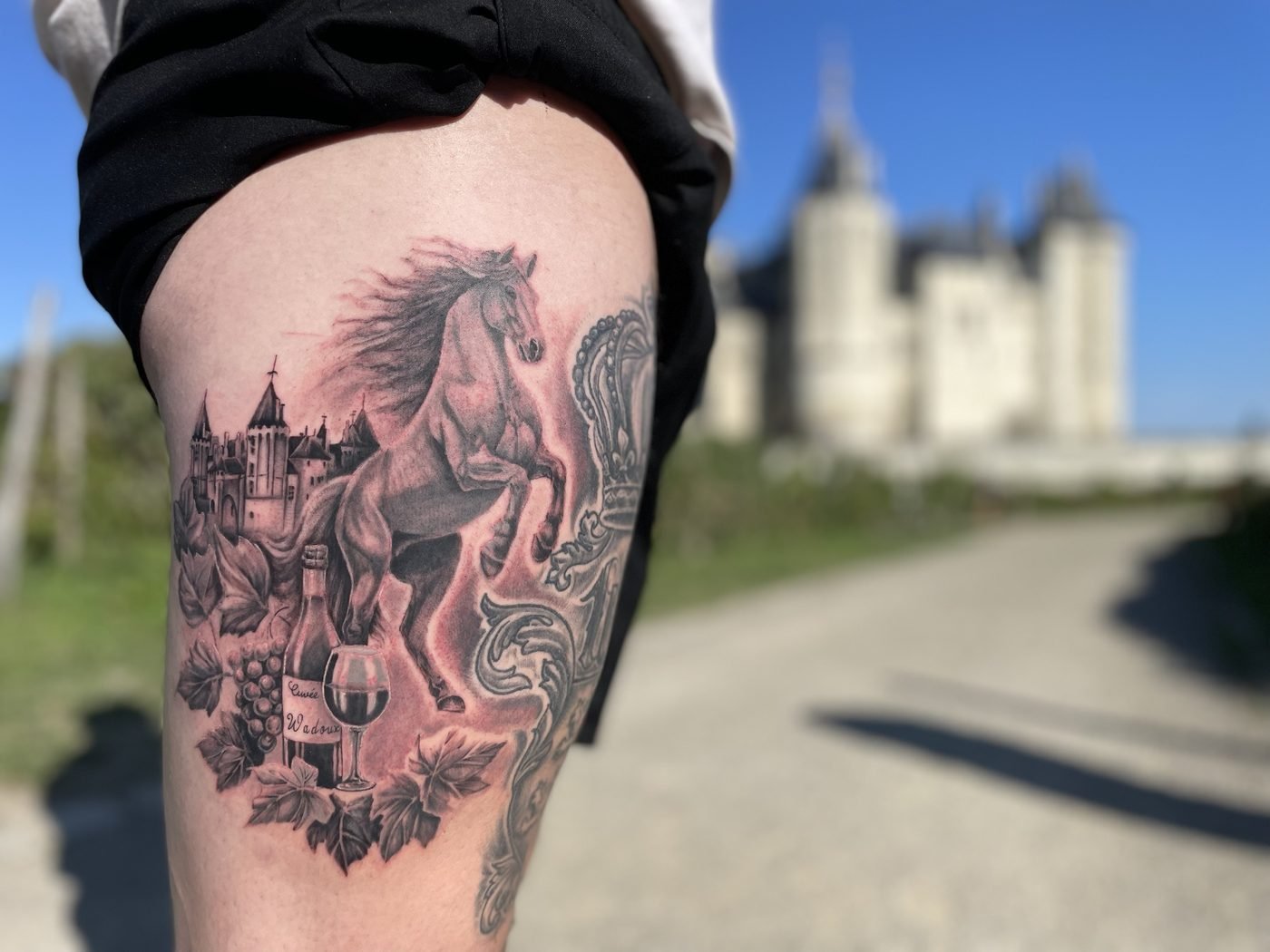 Tatouage réaliste noir et gris : cheval cabré, château de Saumur, vigne et bouteille, sur la cuisse