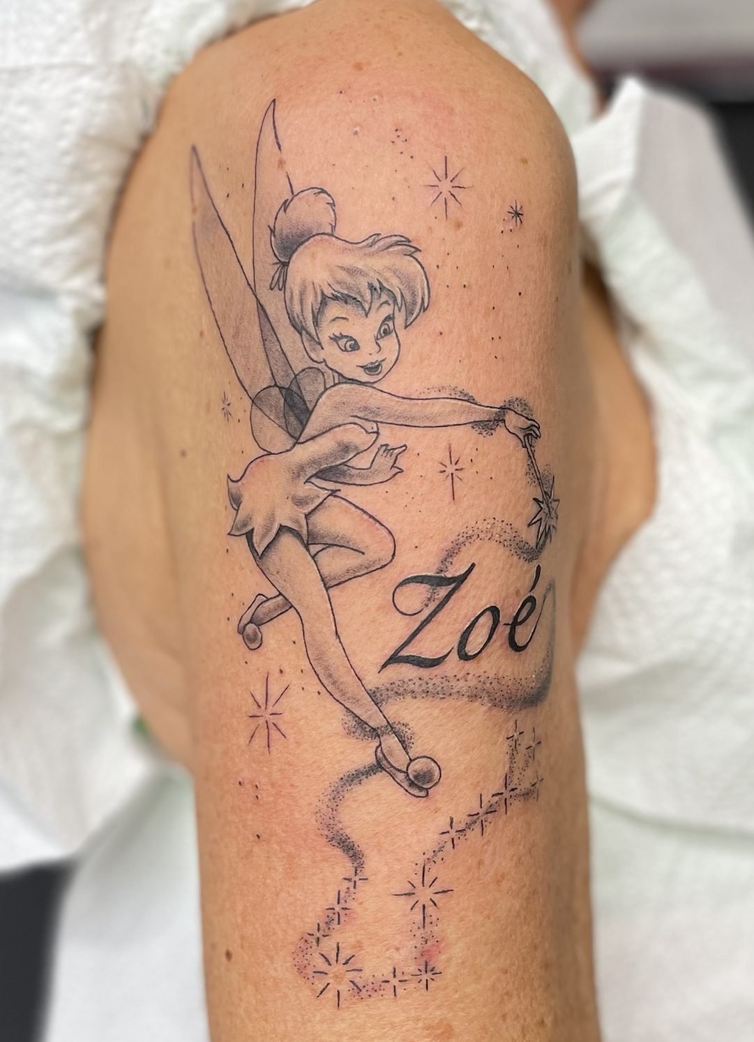 Tatouage de la fée Clochette avec le prénom de sa petite-fille, sur le haut du bras