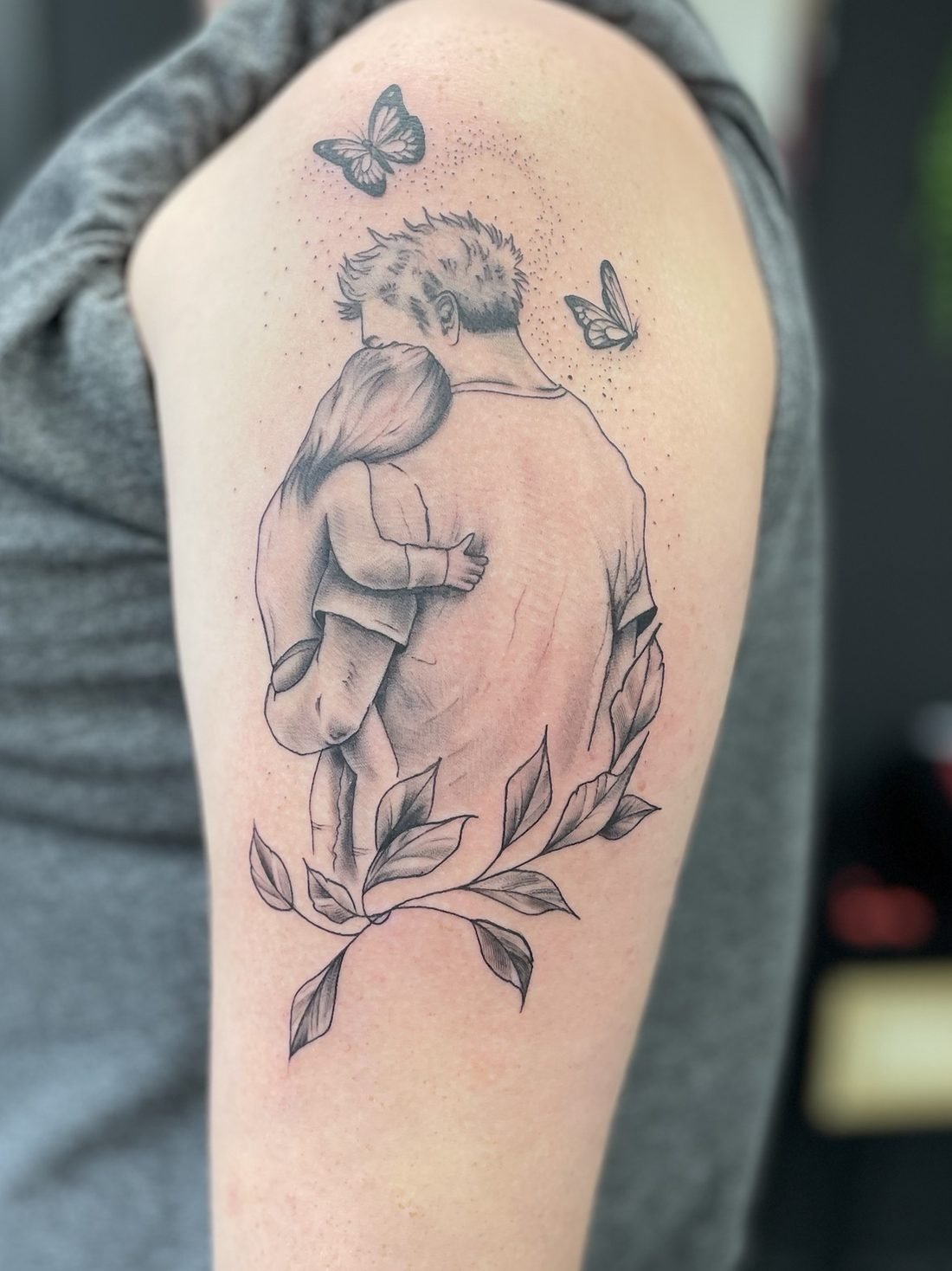 Tatouage hommage : un père tenant son enfant dans ses bras, entouré de papillons et de feuillage