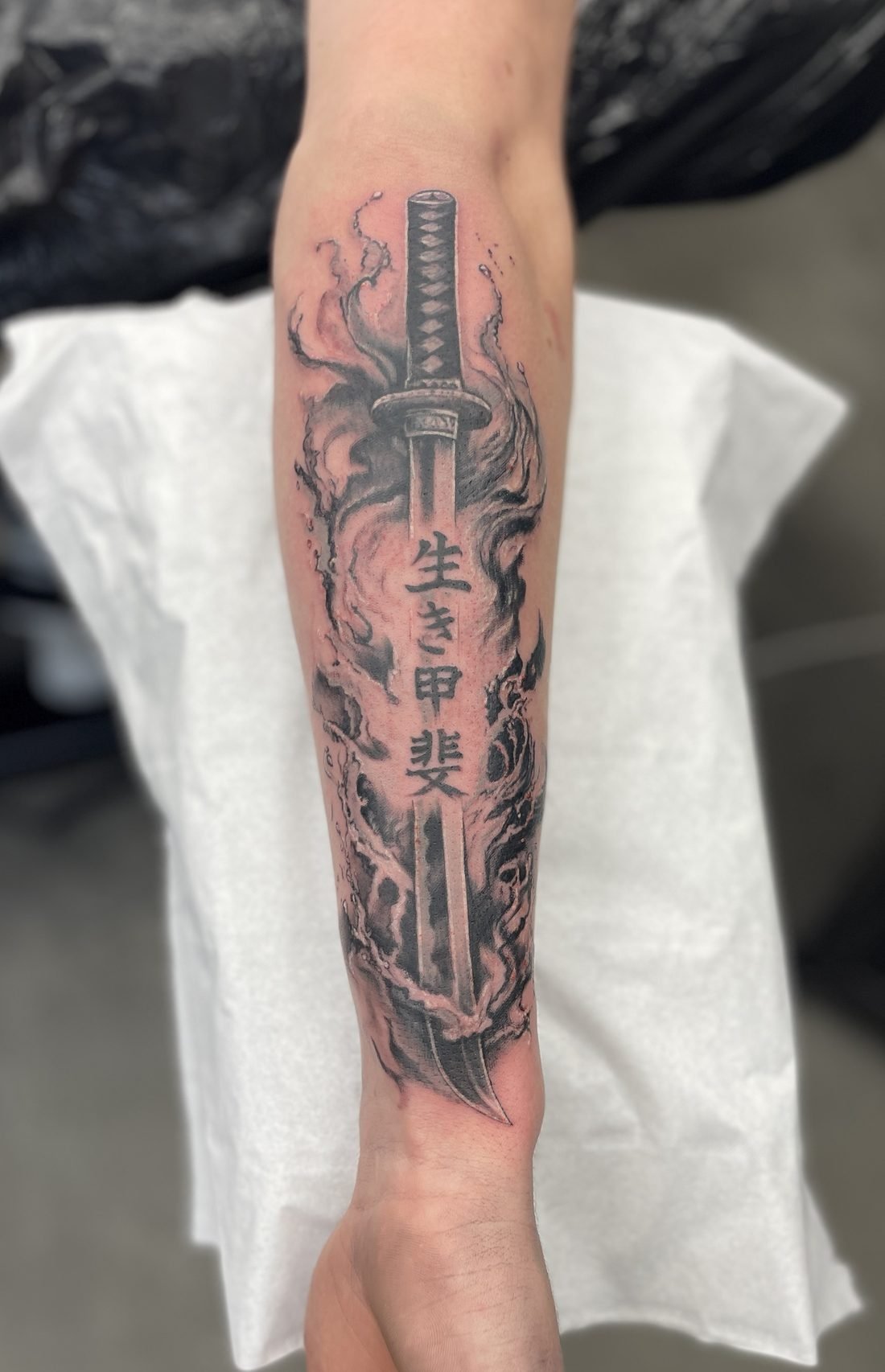 Tatouage noir et gris d’un katana entouré de fumée avec les kanji ikigai, sur l’avant-bras