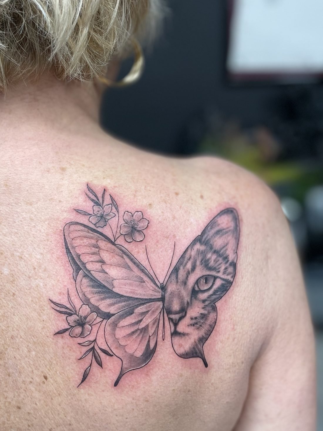 Tatouage noir et gris d’un papillon dont une aile est une tête de chat, avec des fleurs, sur l’omoplate