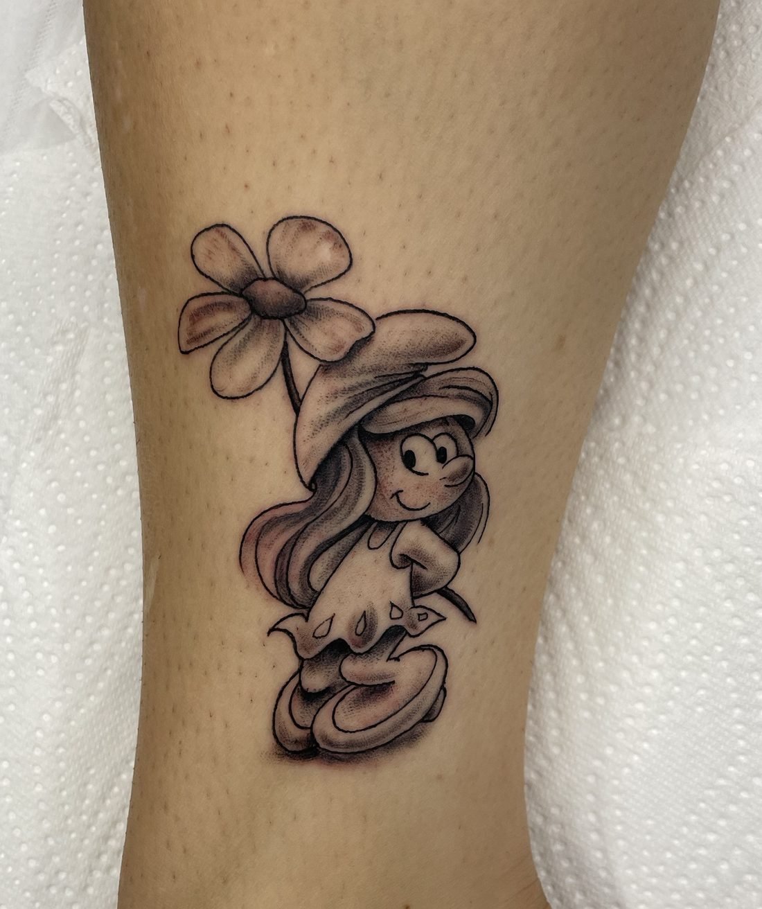 Tatouage noir et gris de la Schtroumpfette tenant une fleur, sur la cheville